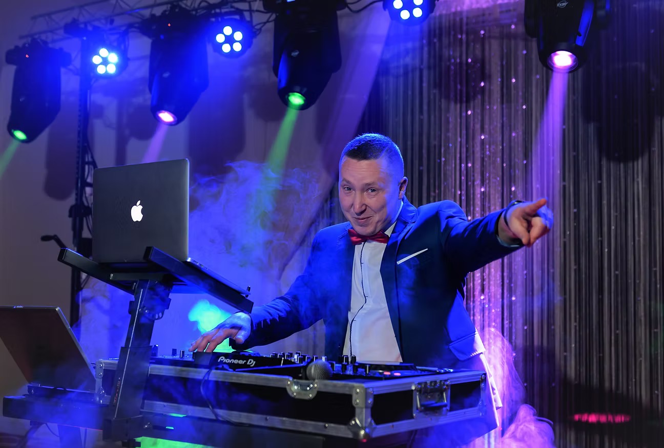Uśmiechnięty DJ w niebieskiej marynarce i czerwonej muszce, wskazujący palcem, grający na sprzęcie Pioneer DJ pod wielobarwnym oświetleniem scenicznym i z laptopem Apple.