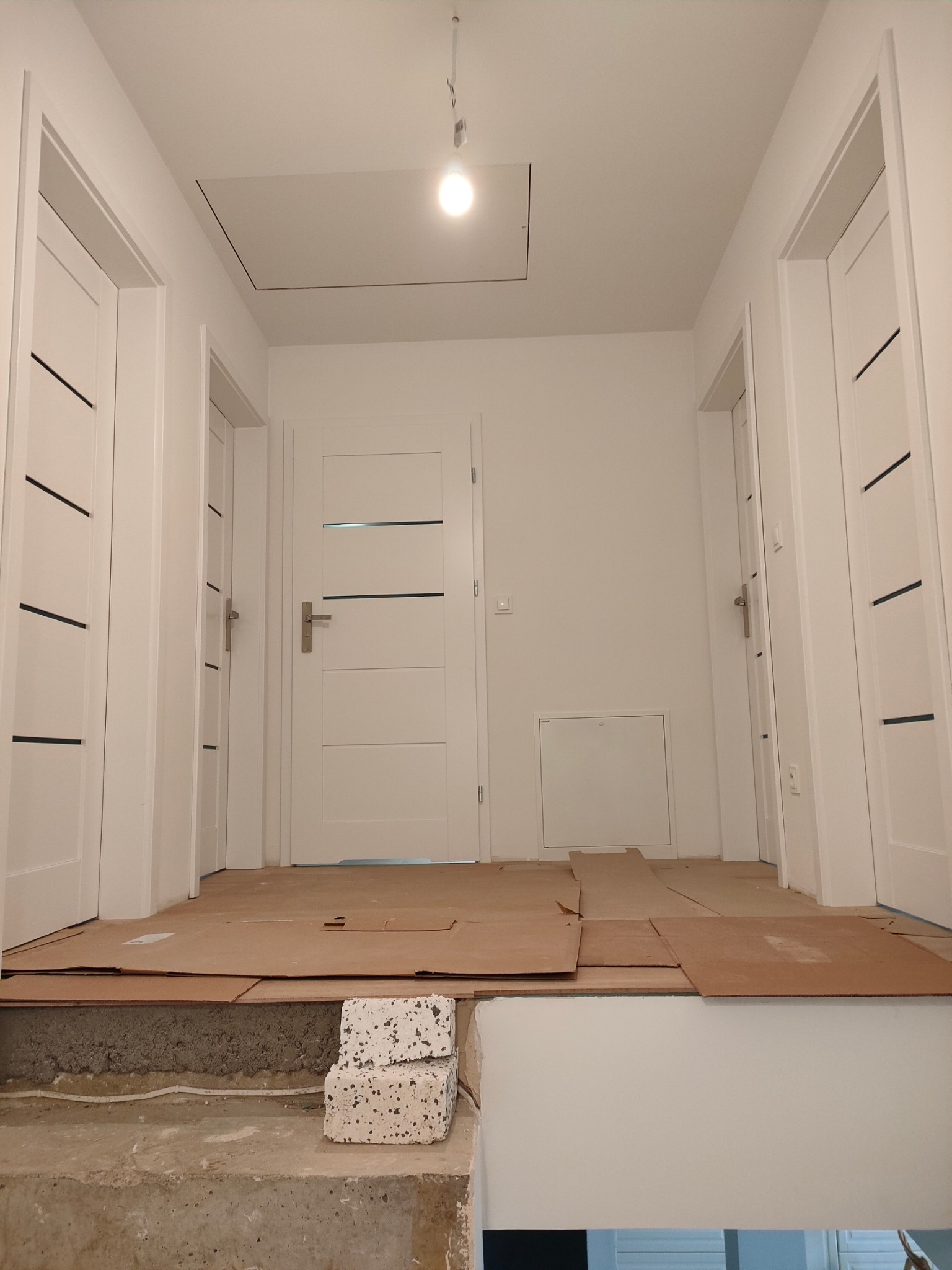 Korytarz w trakcie remontu: białe, nowoczesne drzwi wewnętrzne z poziomymi wstawkami, podłoga zabezpieczona kartonem, widoczne niedokończone schody i cegły.