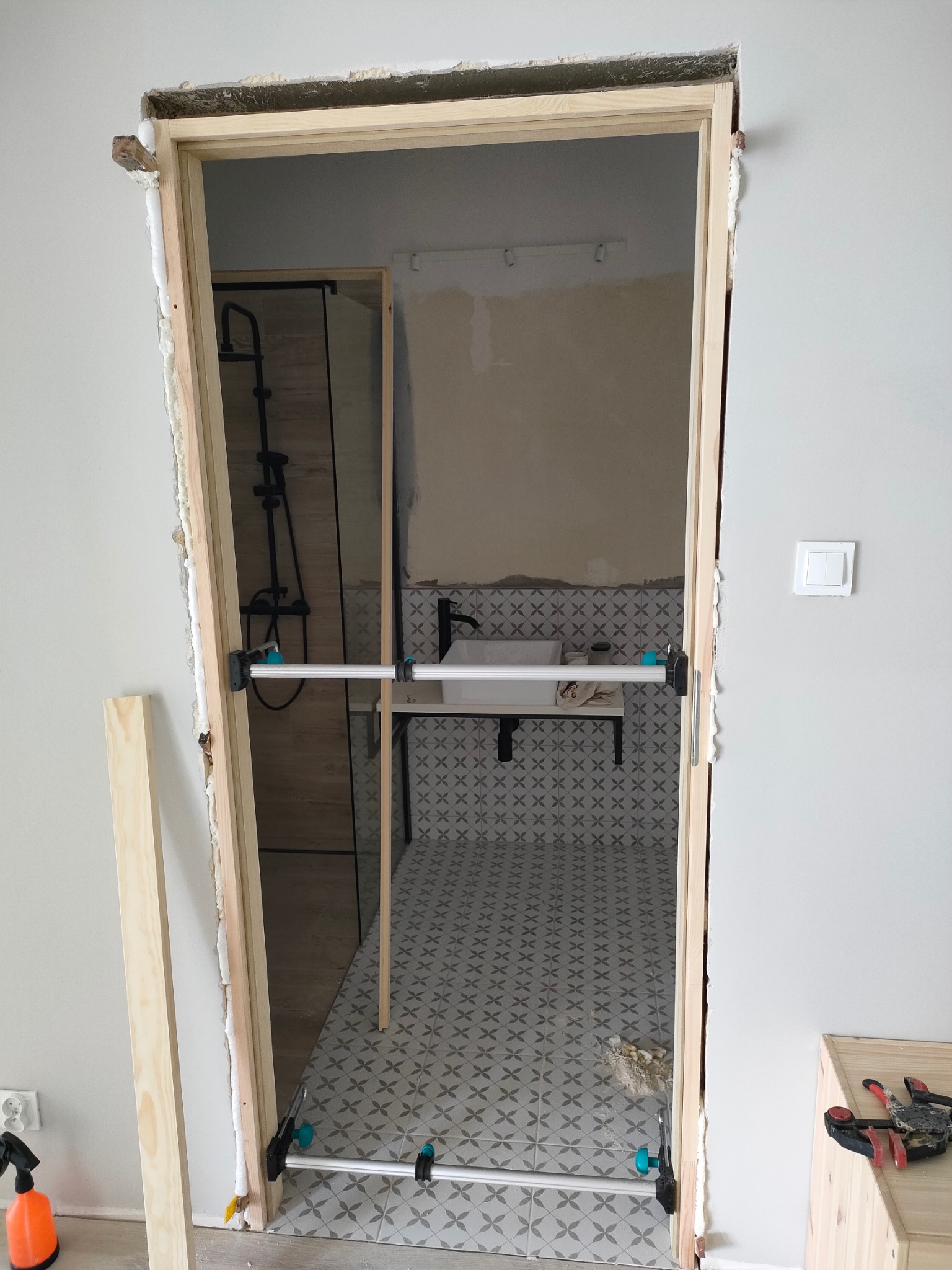 Surowa ościeżnica drewniana, stabilizowana rozpórkami, osadzona w otworze drzwiowym łazienki z widoczną kabiną prysznicową i umywalką na tle wzorzystych płytek podłogowych.