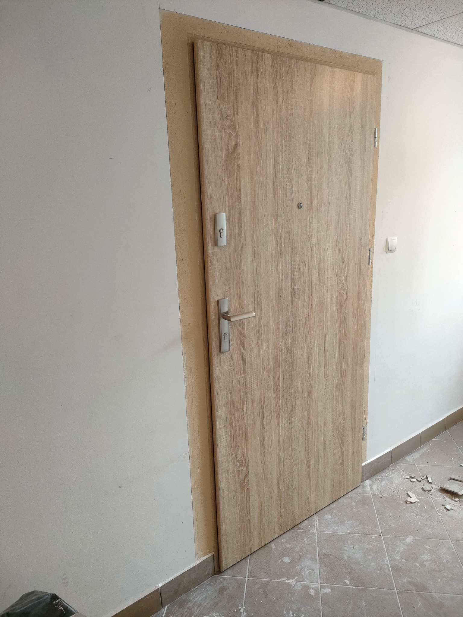 Jasne drewniane drzwi wejściowe, widoczne zamki i klamka, osadzone w ościeżnicy, z widocznymi śladami prac montażowych na podłodze.