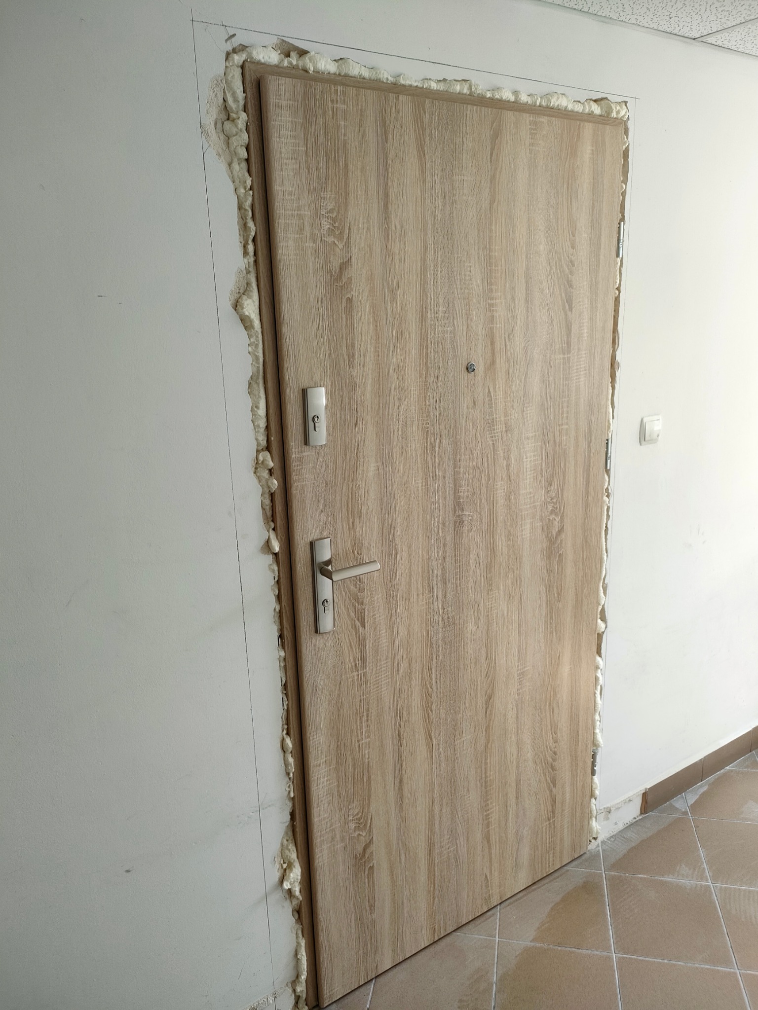 Jasne, drewnopodobne drzwi wejściowe z metalową klamką i wizjerem, osadzone w ościeżnicy wypełnionej pianką montażową, widoczne linie pomocnicze na ścianie.