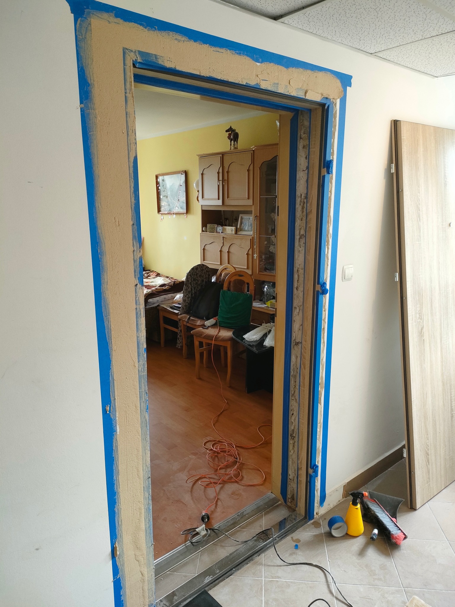 Przygotowanie ościeżnicy do montażu drzwi, z widoczną nieoszlifowaną szpachlą i taśmą malarską, w tle widoczny pokój z meblami i otwarte drzwi.