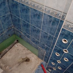 -uBracii- - Zdemontowana kabina prysznicowa z widocznymi niebieskimi płytkami, rurą odpływową i poziomicą. Ślady po demontażu na podłodze i ścianach.