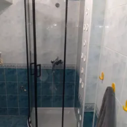 Nowoczesna kabina prysznicowa z czarną ramą w wykafelkowanej łazience. Widoczny panel prysznicowy i szary ręcznik na żółtym haczyku. Ujęcie z bliska.