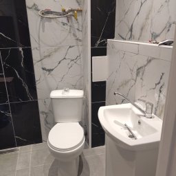 -uBracii- - Wykończona łazienka z białą armaturą i czarno-białymi płytkami imitującymi marmur. Widoczna umywalka z szafką, toaleta i fragment instalacji hydraulicznej na ścianie.