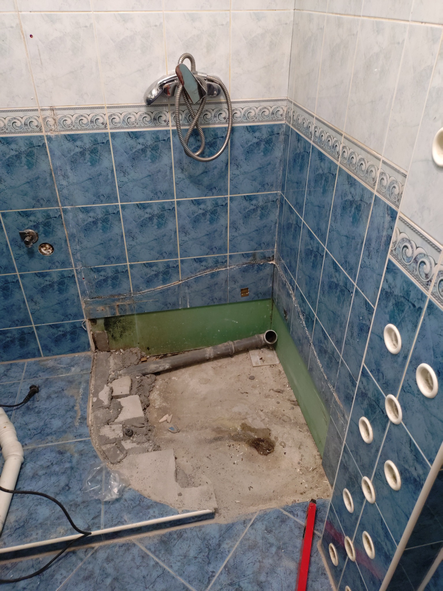 Łazienka w trakcie remontu: zdemontowana kabina prysznicowa, widoczne rury odpływowe i fragmenty uszkodzonych płytek ściennych w odcieniach błękitu i bieli.