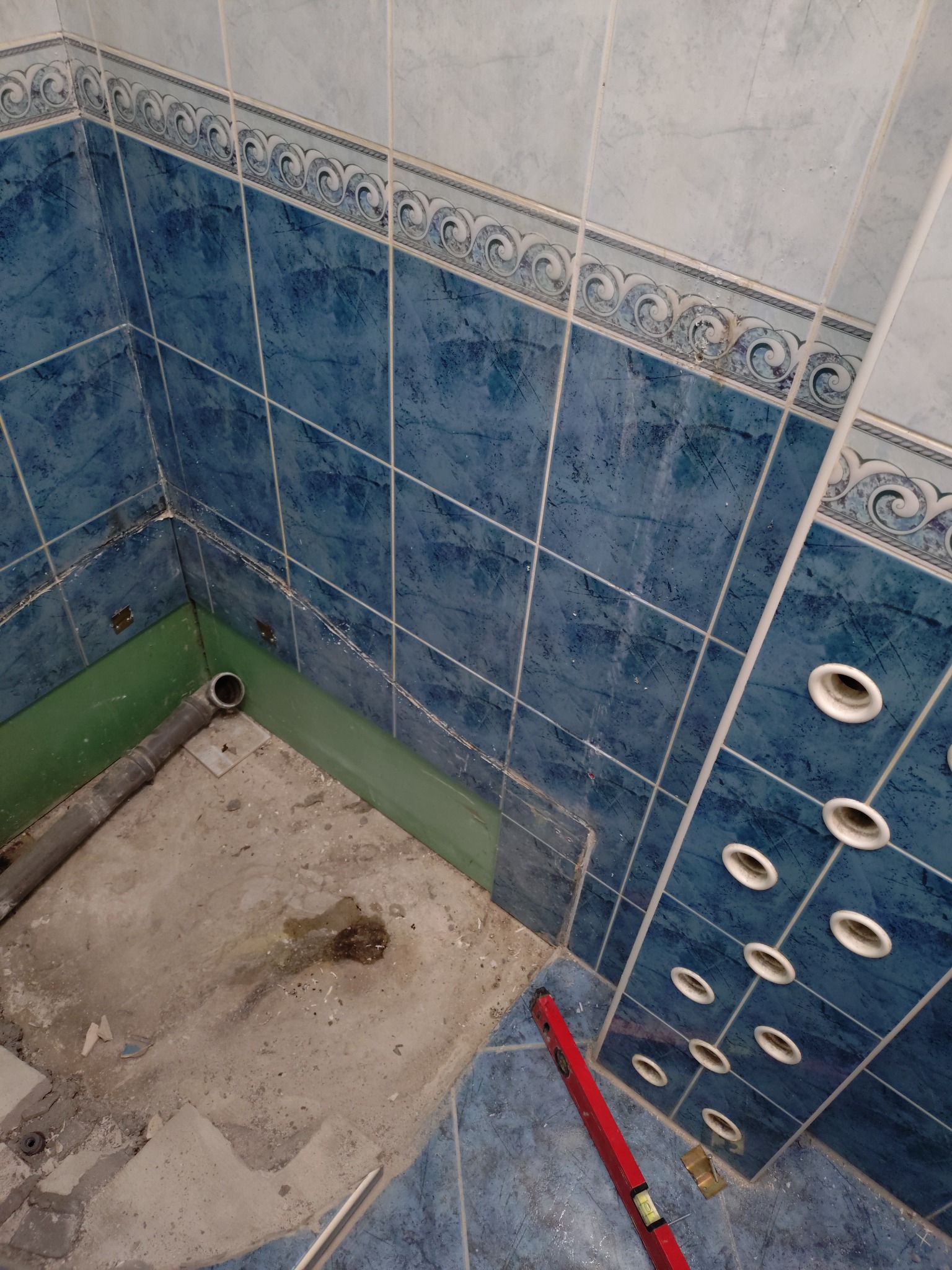 Zdemontowana kabina prysznicowa z widocznymi niebieskimi płytkami, rurą odpływową i poziomicą. Ślady po demontażu na podłodze i ścianach.