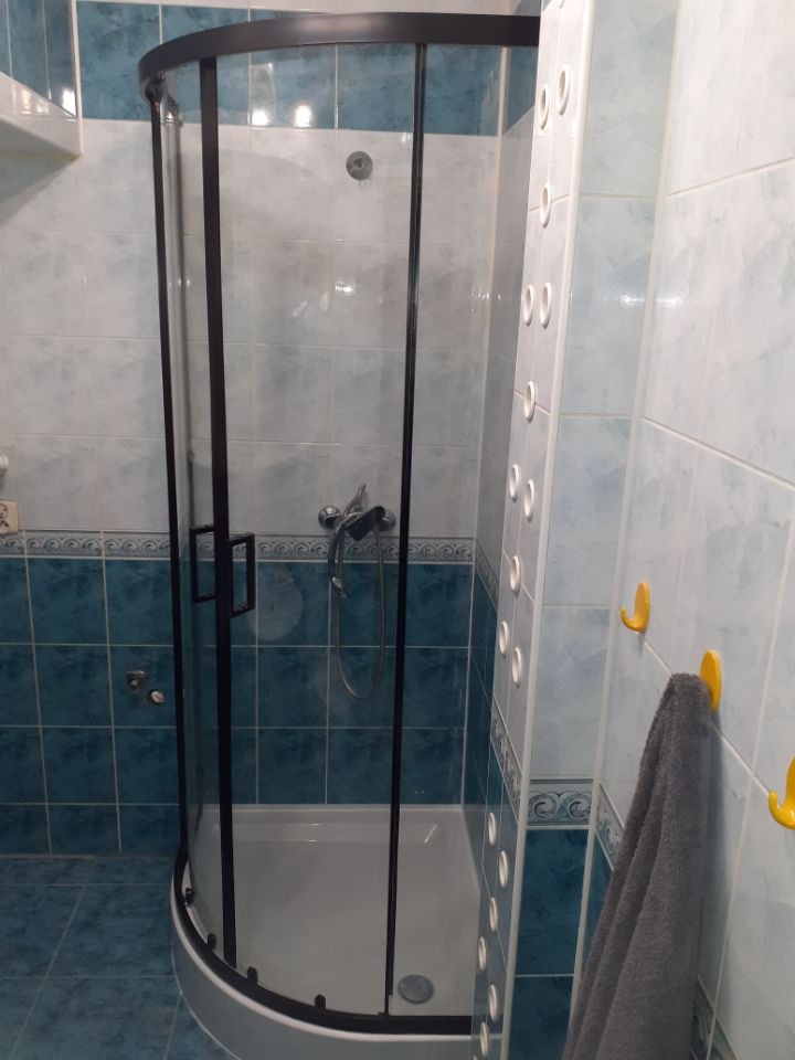 Nowoczesna kabina prysznicowa z czarną ramą w wykafelkowanej łazience. Widoczny panel prysznicowy i szary ręcznik na żółtym haczyku. Ujęcie z bliska.