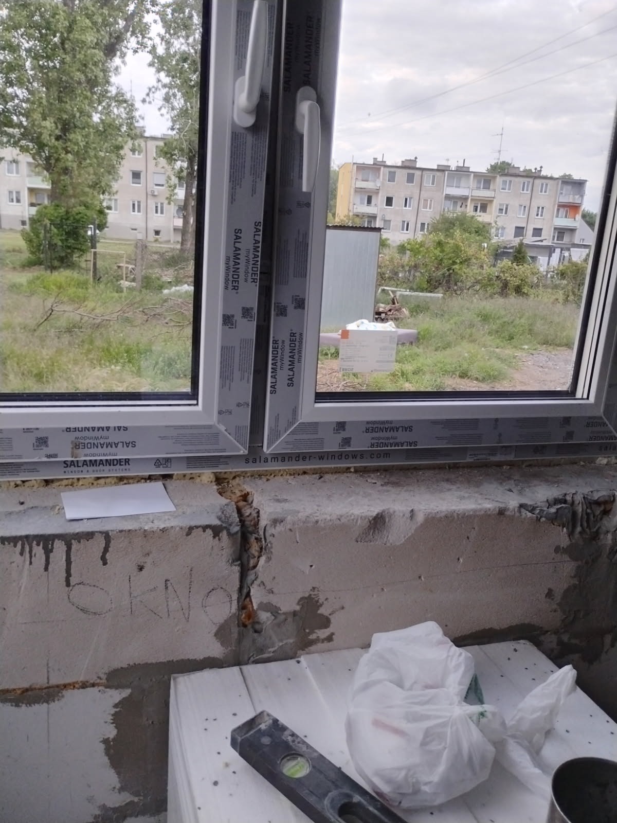 Nowe okno Salamander z widokiem na budynek mieszkalny i zieleń. W tle nieotynkowana ściana z poziomnicą i napisem 'OKNO'.