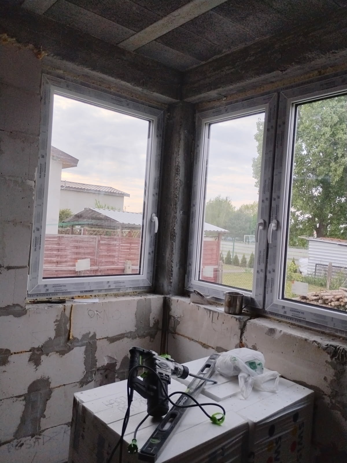Montaż dwóch okien Salamander w surowym wnętrzu, widoczne narzędzia i bloczki betonowe. Widok na zewnątrz, dom i drzewa.
