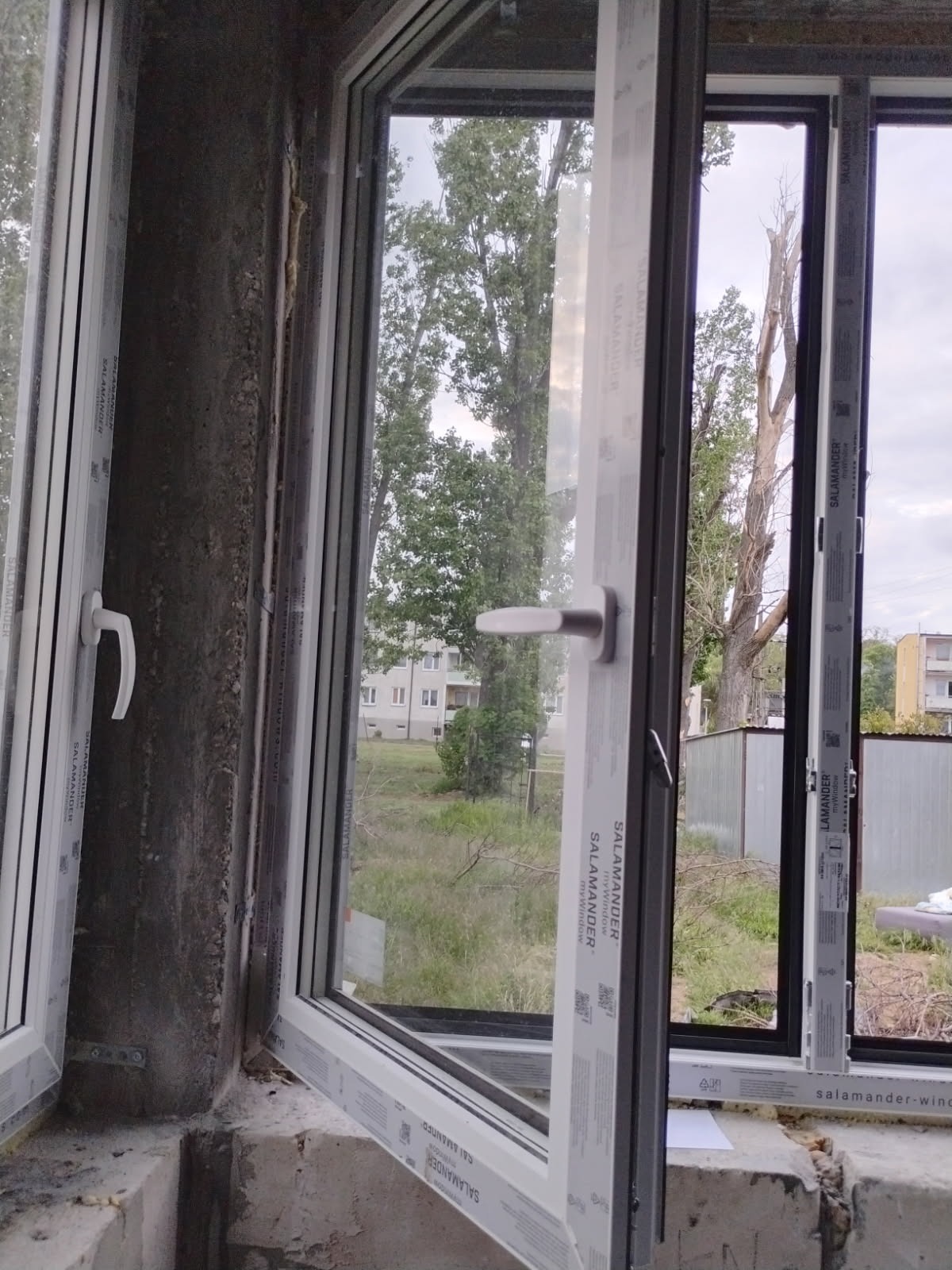 Montaż okna Salamander w surowym stanie, widok na zewnątrz. Okno otwarte, z widoczną klamką. Tło: budynek i zieleń. Surowy mur z widoczną zaprawą.