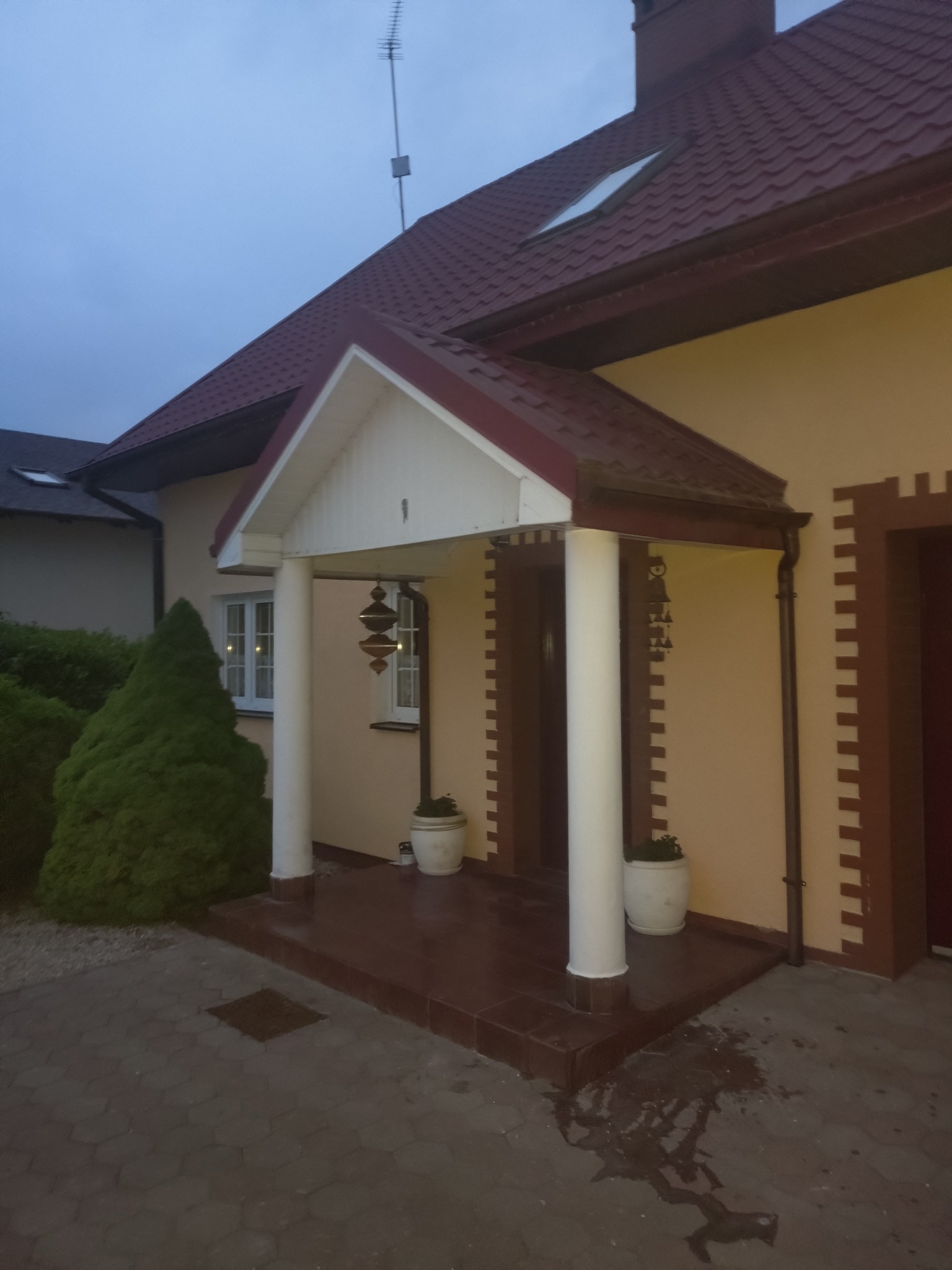 Elewacja domu jednorodzinnego po malowaniu na jasny kolor, widoczny fragment dachu z dachówki w odcieniach czerwieni, dekoracyjne wykończenia okien i drzwi.