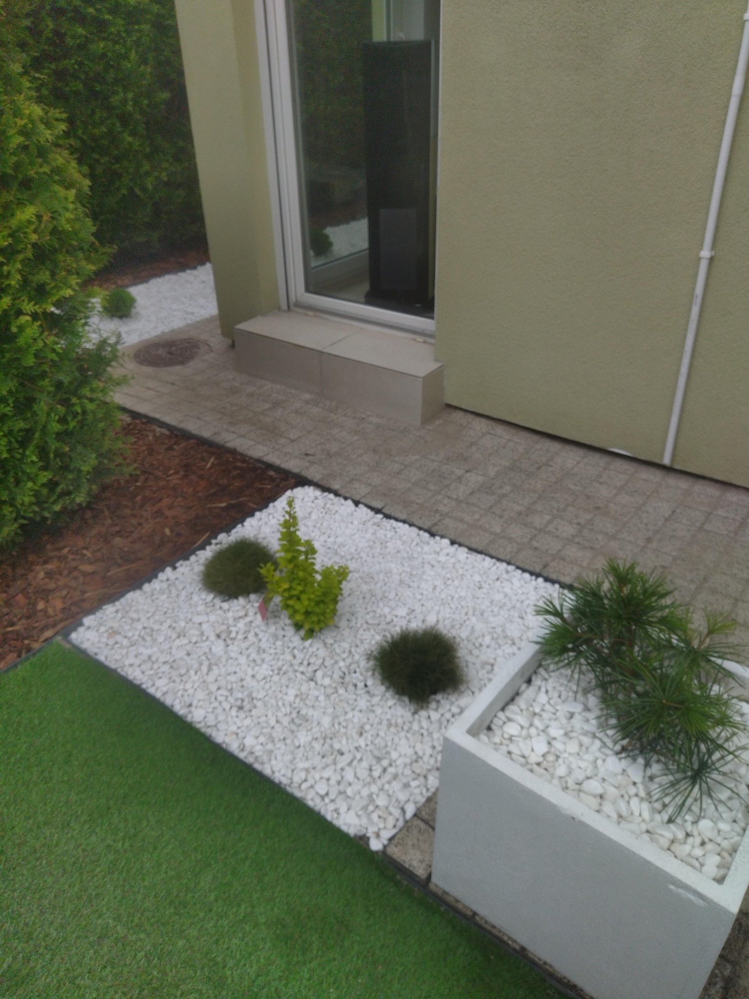 Aranżacja ogrodu przy domu z białymi kamieniami, sztuczną trawą i roślinami w donicach.