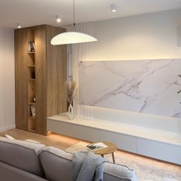 Dmytro Tytorenko - Nowoczesny salon z drewnianą szafą, białą szafką RTV z marmurowym panelem i rośliną w donicy. Minimalistyczny design, ciepłe oświetlenie.
