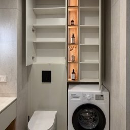 Dmytro Tytorenko - Biała szafa na wymiar w łazience z oświetleniem LED, zabudowana pralka i toaleta podwieszana. Minimalistyczny design, szare płytki podłogowe i ścienne.