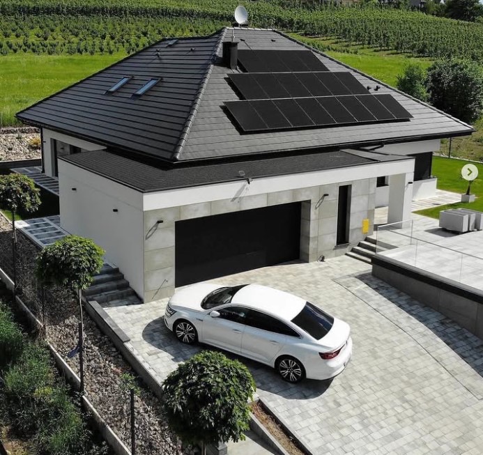 Nowoczesny dom jednorodzinny z czarnym dachem i panelami słonecznymi, biała elewacja, garaż z czarną bramą, podjazd z kostki brukowej, biały samochód osobowy, w tle zielony krajobraz.