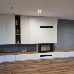 Ma&Ja Meble - Nowoczesna zabudowa ściany z szafkami, półkami i kominkiem, białe fronty z drewnianymi akcentami, szara ściana w tle, drewniana podłoga.