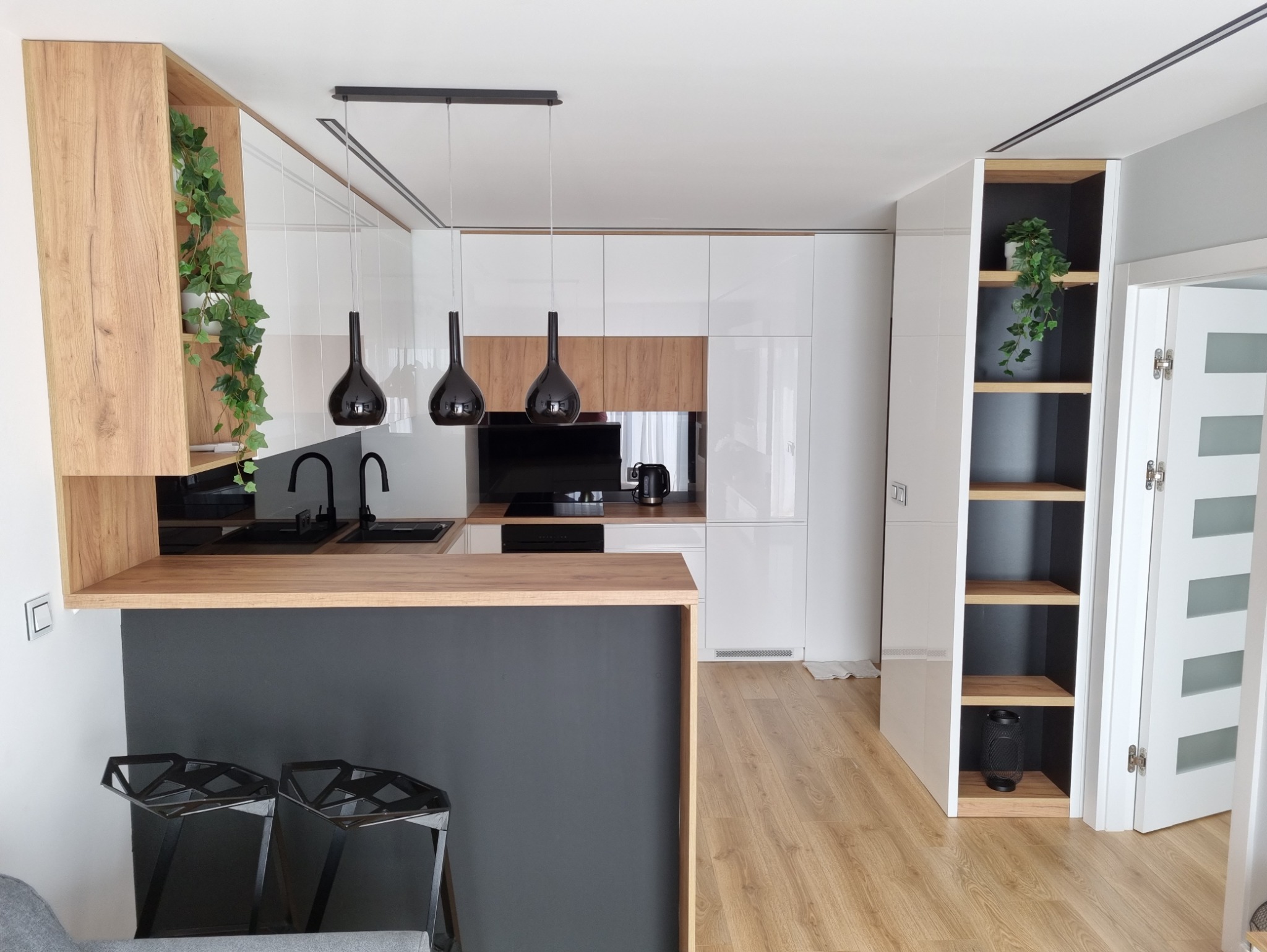 Nowoczesna kuchnia z białymi, lakierowanymi frontami, drewnianymi akcentami, czarnymi lampami wiszącymi i blatem z barkiem, uzupełniona o otwarty regał z dekoracyjną rośliną.
