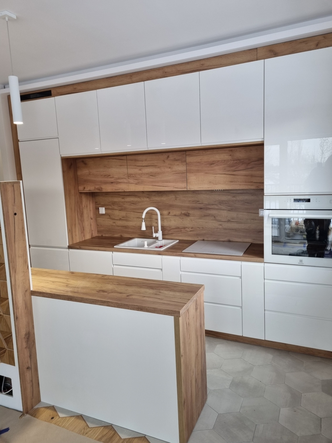 Nowoczesna kuchnia z białymi, lakierowanymi frontami i elementami z drewna dębowego, wyspa kuchenna z blatem drewnianym, heksagonalna podłoga.