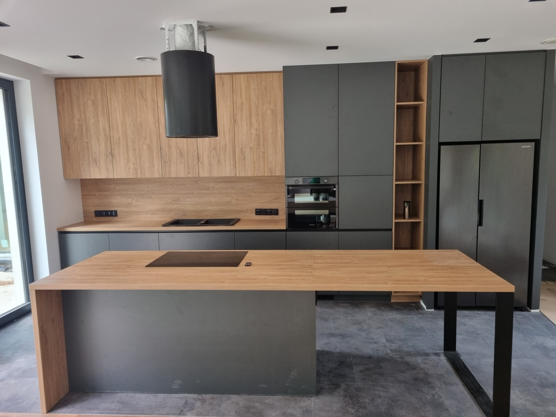 Nowoczesna kuchnia z wyspą, fronty drewnopodobne i szare, czarny okrągły okap, płyta indukcyjna na wyspie, szare płytki na podłodze.