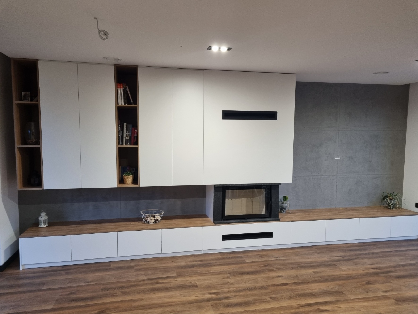 Nowoczesna zabudowa ściany z szafkami, półkami i kominkiem, białe fronty z drewnianymi akcentami, szara ściana w tle, drewniana podłoga.