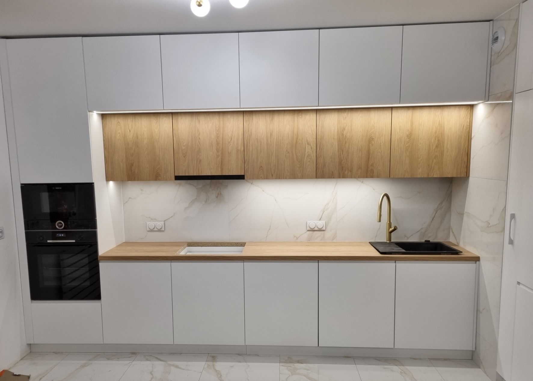 Nowoczesna kuchnia z białymi szafkami dolnymi i górnymi w kolorze jasnego drewna, złota bateria, marmurowe płytki na ścianie, zabudowany piekarnik Bosch.