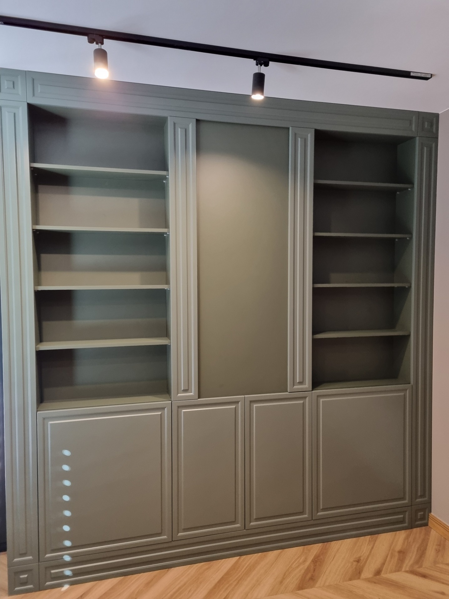 Duża, grafitowa szafa wnękowa z otwartymi półkami i zamkniętymi szafkami, oświetlona dwoma reflektorami sufitowymi, na podłodze parkiet w jodełkę.