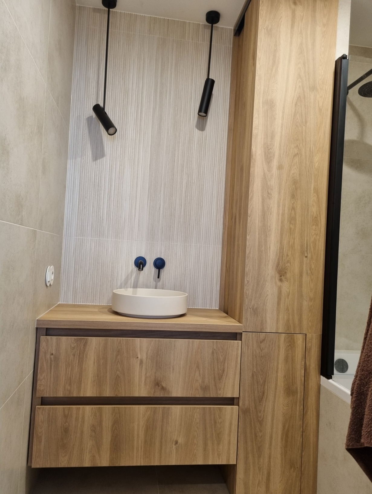 Nowoczesna łazienka z szafką pod umywalką w odcieniach drewna, okrągłą białą umywalką nablatową i dwoma czarnymi lampami sufitowymi.