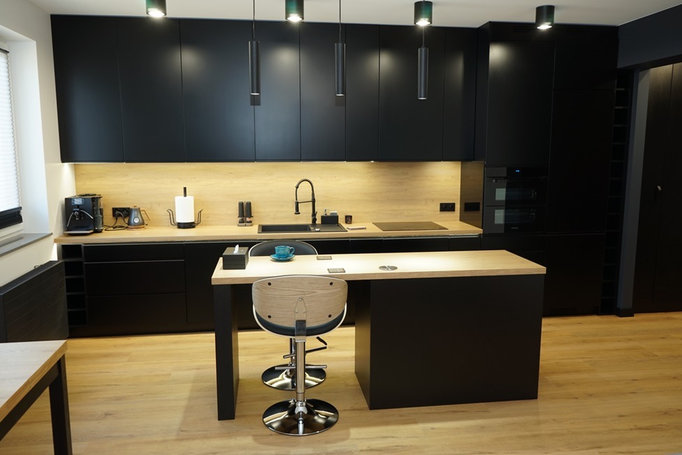 Nowoczesna, czarna kuchnia z drewnianym blatem, wyspą kuchenną i hokerami. Oświetlenie punktowe nad blatem i wyspą. Na blacie ekspres do kawy, czajnik i akcesoria kuchenne.