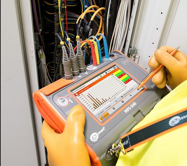 Miernik MPI-540 w dłoni elektryka, analiza wykresów harmonicznych na tle otwartej szafy z kablami. Pomiar parametrów instalacji elektrycznej.