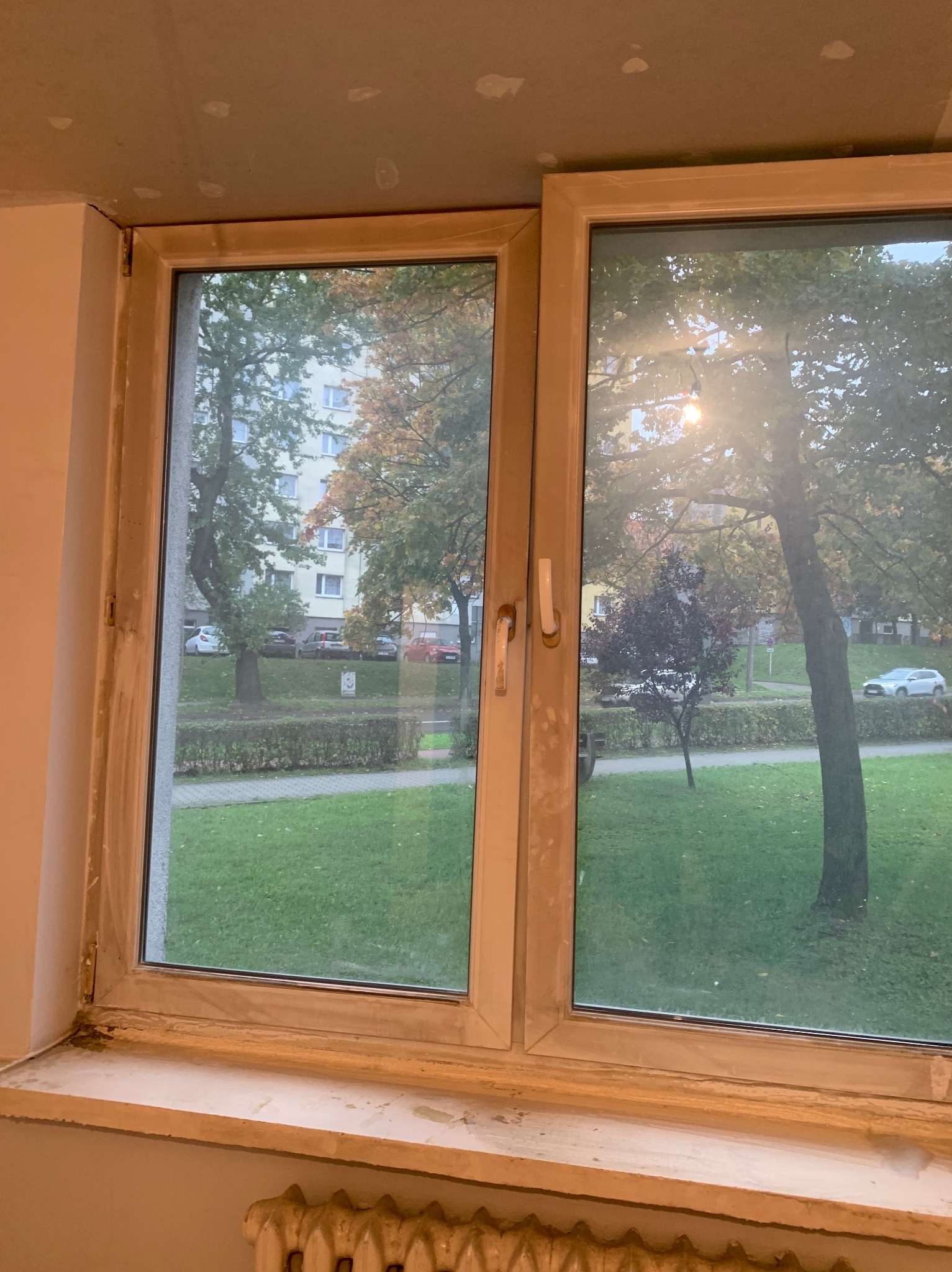 Okna PCV po montażu, widoczne ślady po pracach wykończeniowych. Na zewnątrz zieleń, budynki mieszkalne i jesienne drzewa w tle.