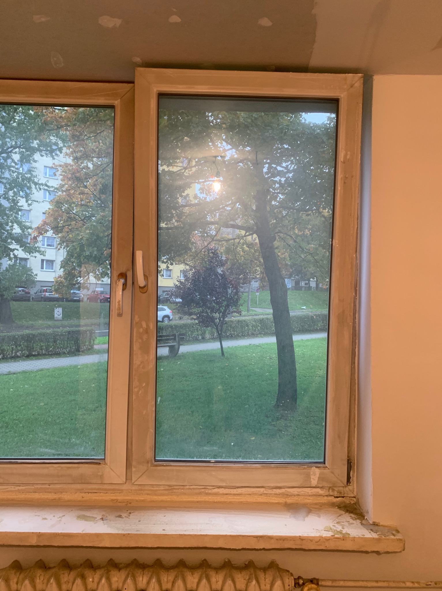 Okna PCV po montażu, widoczne ślady po remoncie na parapecie i ścianie. Za oknem widok na drzewa i blok mieszkalny.