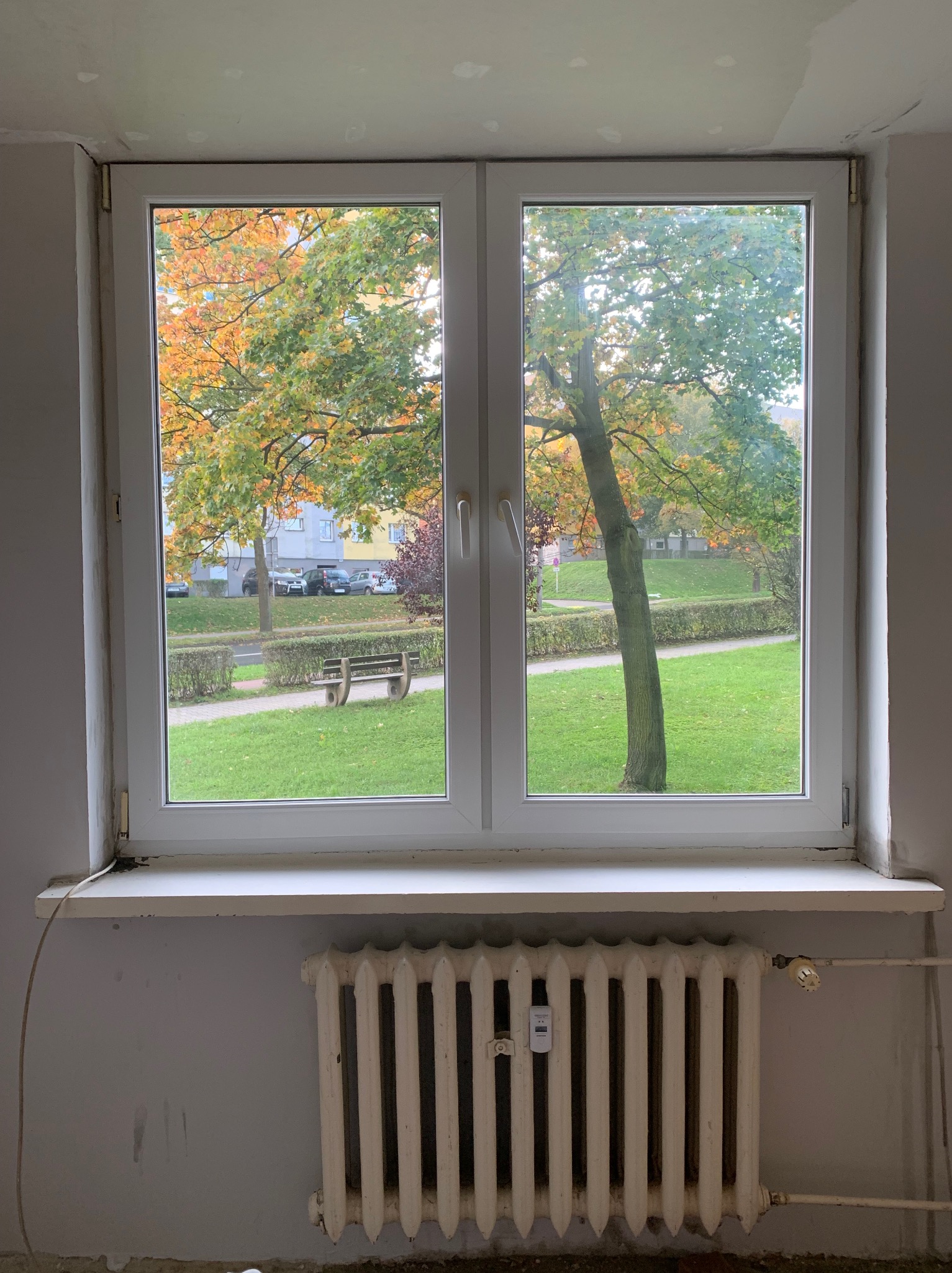 Białe okno z widokiem na park jesienią. Szary pokój z grzejnikiem, ślady po remoncie na ścianach. Pod parapetem widoczny kabel.