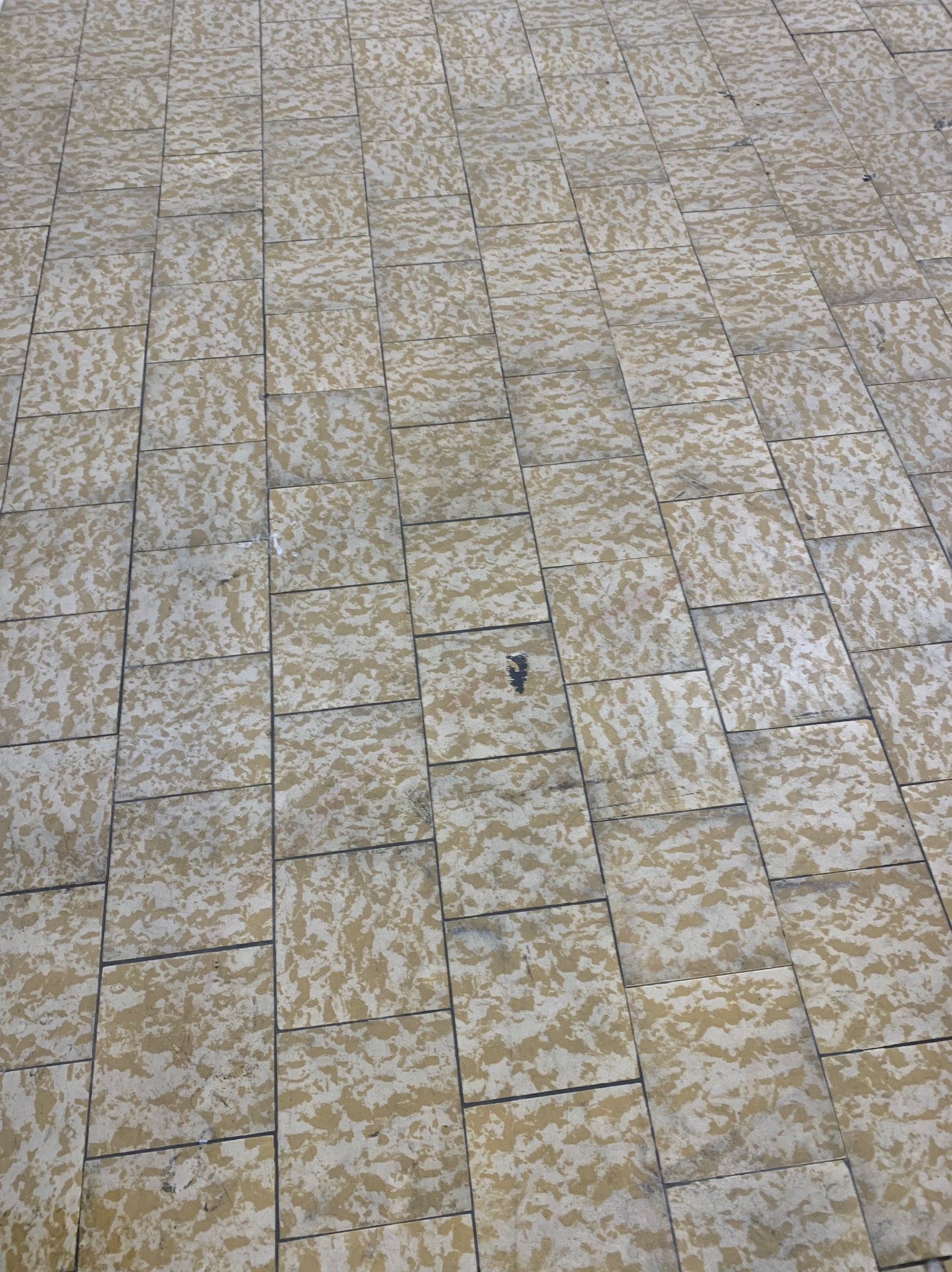 Ułożona kostka brukowa w odcieniach beżu i szarości, widoczna z góry pod kątem. Na powierzchni widoczne zabrudzenia, w jednym miejscu ciemna plama.