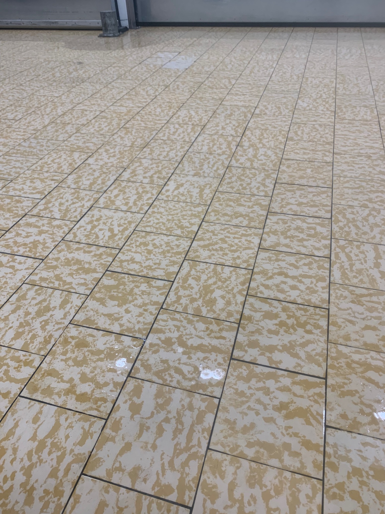 Błyszcząca, świeżo umyta podłoga z płytek w odcieniach beżu i brązu, z ciemnymi fugami, odbijająca światło, sugerująca niedawne sprzątanie w przestrzeni komercyjnej.