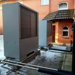 Dwie szare pompy ciepła zamontowane na zewnątrz budynku w Toruniu. Widoczne rury i elementy instalacji na tle ceglanej ściany i lekko ośnieżonego terenu.