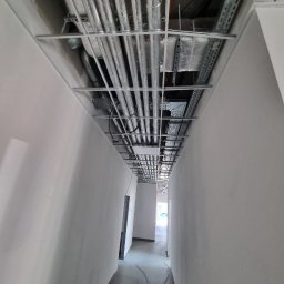 INTEC INSTALACJE TECHNICZNE SPÓŁKA Z OGRANICZONĄ ODPOWIEDZIALNOŚCIĄ - Instalacje wentylacyjne i rury pod sufitem w korytarzu, widok z dołu. Równoległe rury w srebrnej izolacji, metalowe korytka kablowe, surowy stan wykończenia.