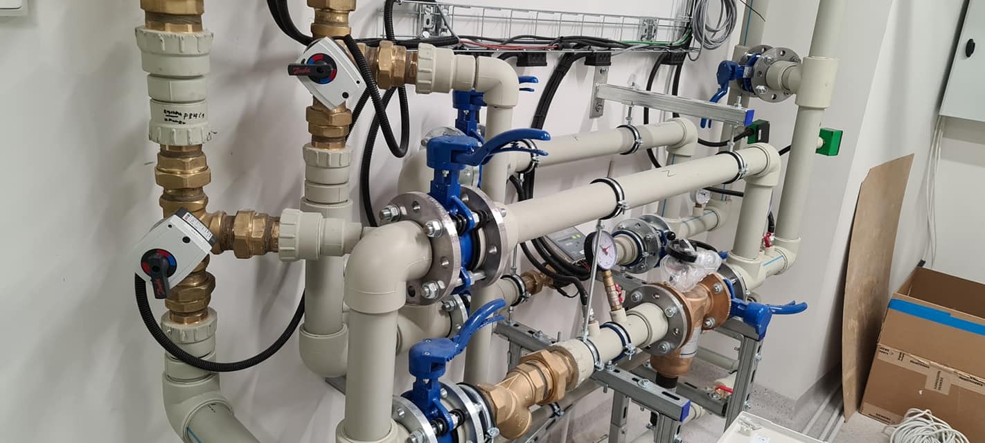 Złożona instalacja hydrauliczna z rur z tworzywa i mosiądzu, zaworami z niebieskimi uchwytami i manometrem, zamontowana na białej ścianie z okablowaniem.