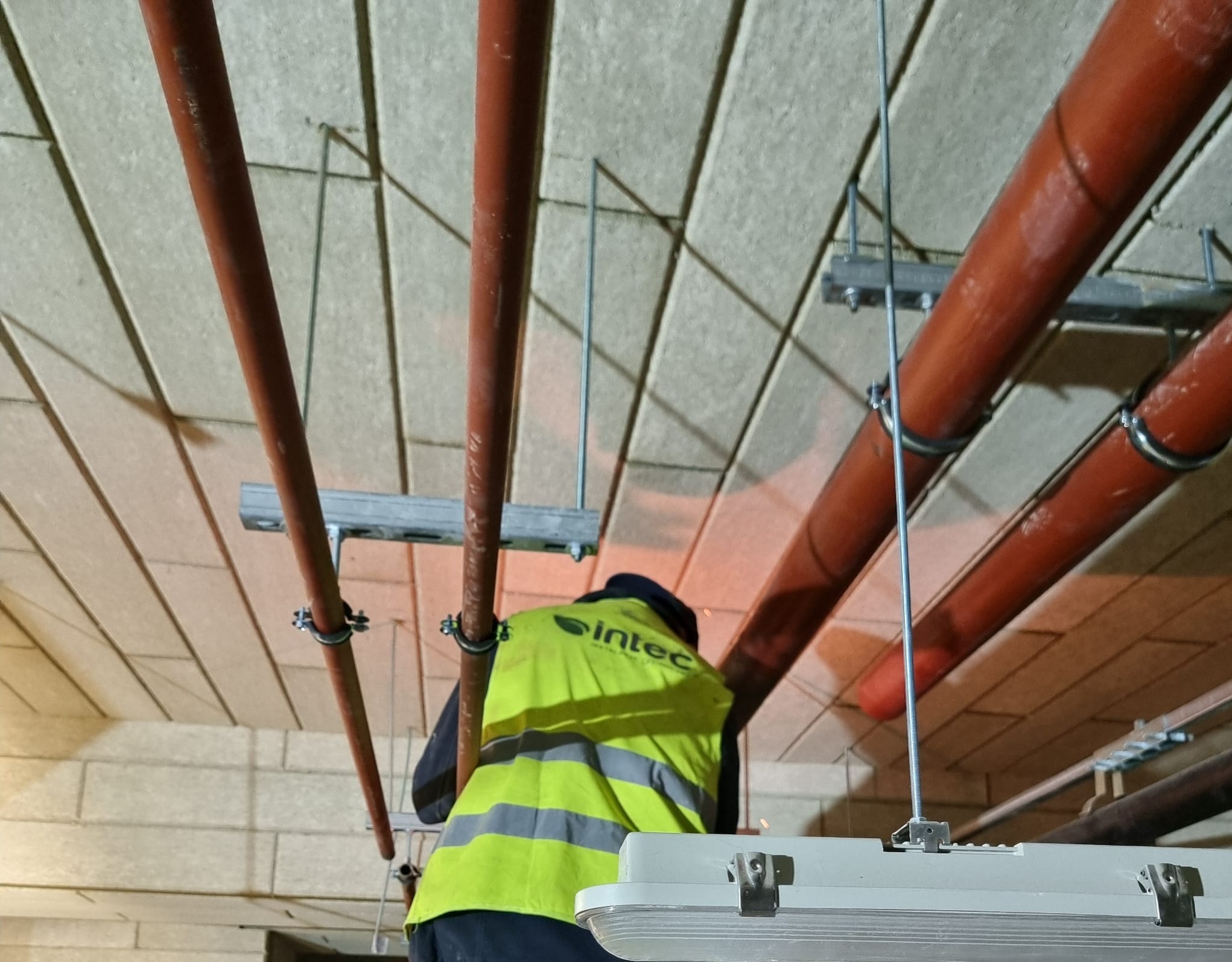 Montaż instalacji: czerwone rury pod sufitem, mocowane metalowymi uchwytami. Pracownik w żółtej kamizelce montuje oświetlenie. Widok z dołu.