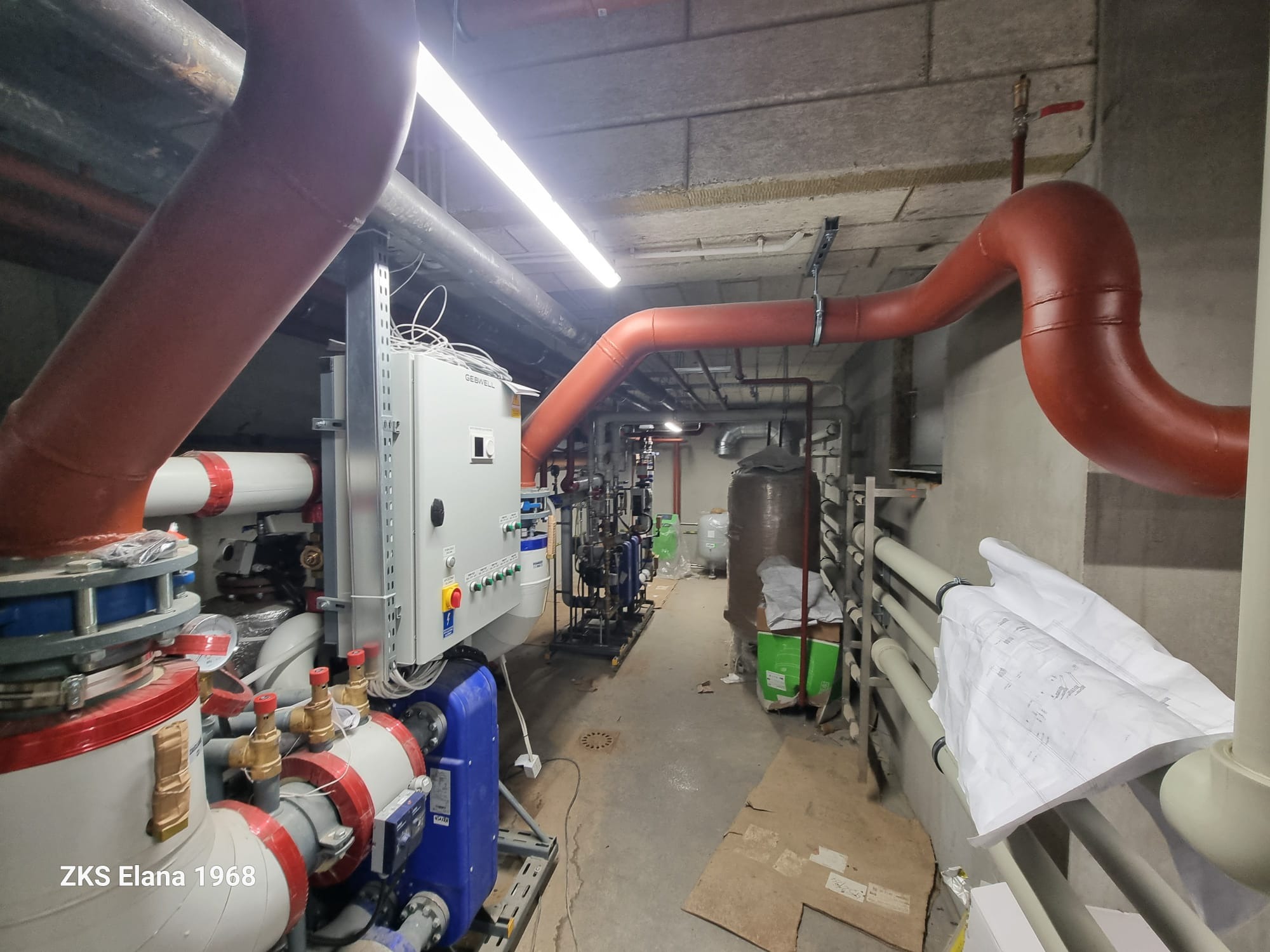 Węzeł cieplny w piwnicy: rozbudowany system rur w kolorze czerwonym i szarym, podłączony do centrali sterującej. Widoczne plany instalacji i elementy wyposażenia.