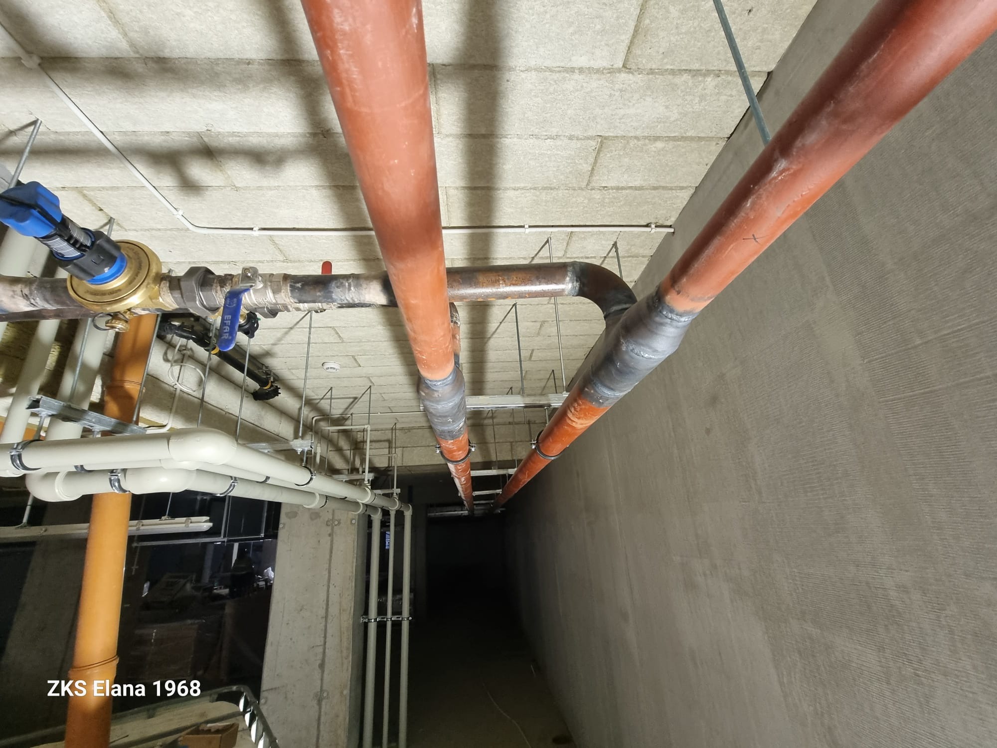 Instalacja rur w piwnicy: pomarańczowe i białe rury pod sufitem, widoczne złącza i zawory. Perspektywa od dołu, surowy beton.