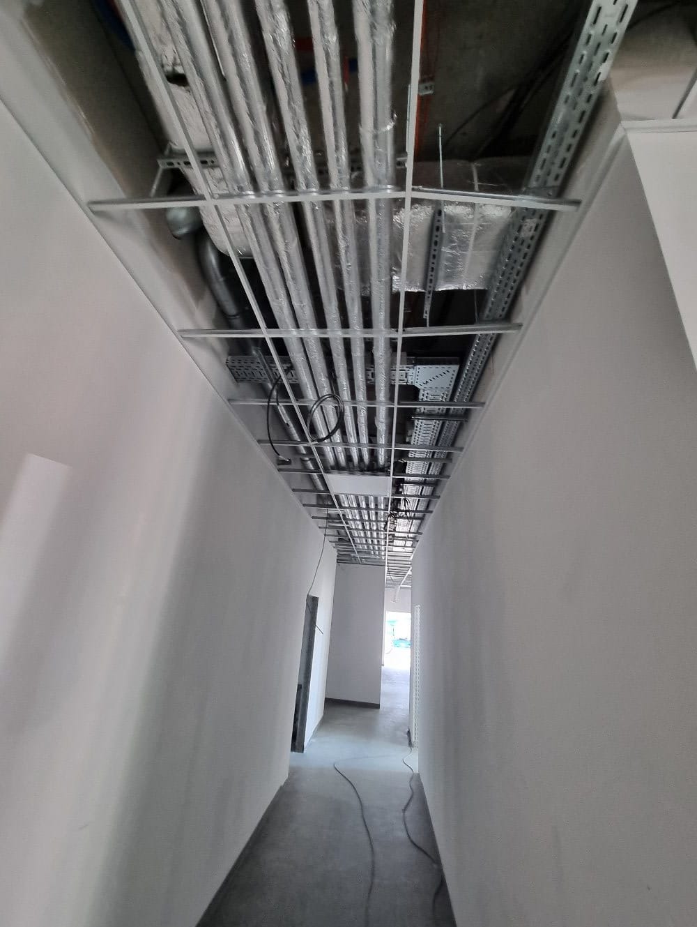 Instalacje wentylacyjne i rury pod sufitem w korytarzu, widok z dołu. Równoległe rury w srebrnej izolacji, metalowe korytka kablowe, surowy stan wykończenia.