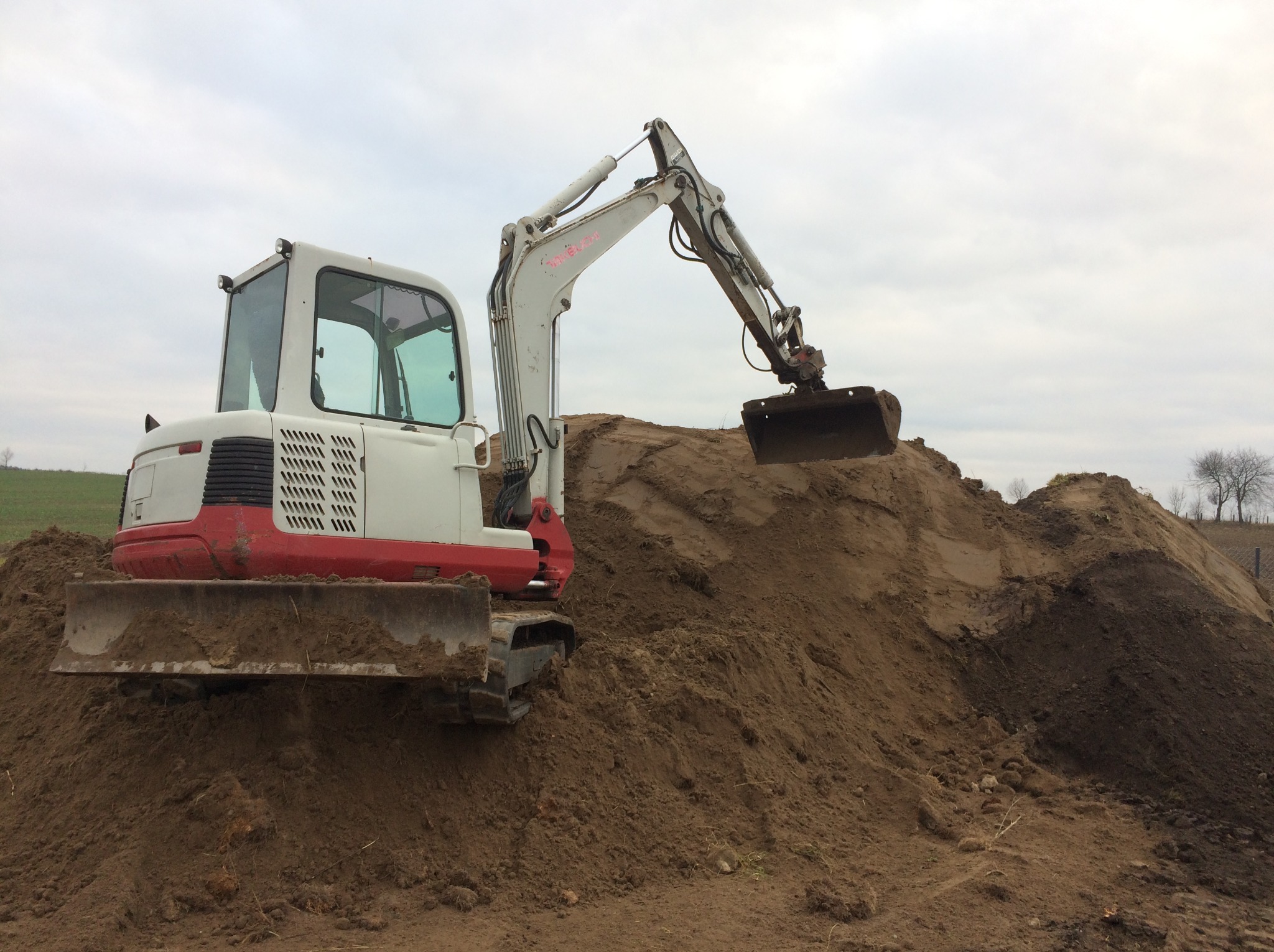 Biało-czerwona mini koparka Takeuchi na gąsienicach, z łyżką skierowaną w dół, pracuje na usypanej górze ziemi w pochmurny dzień.