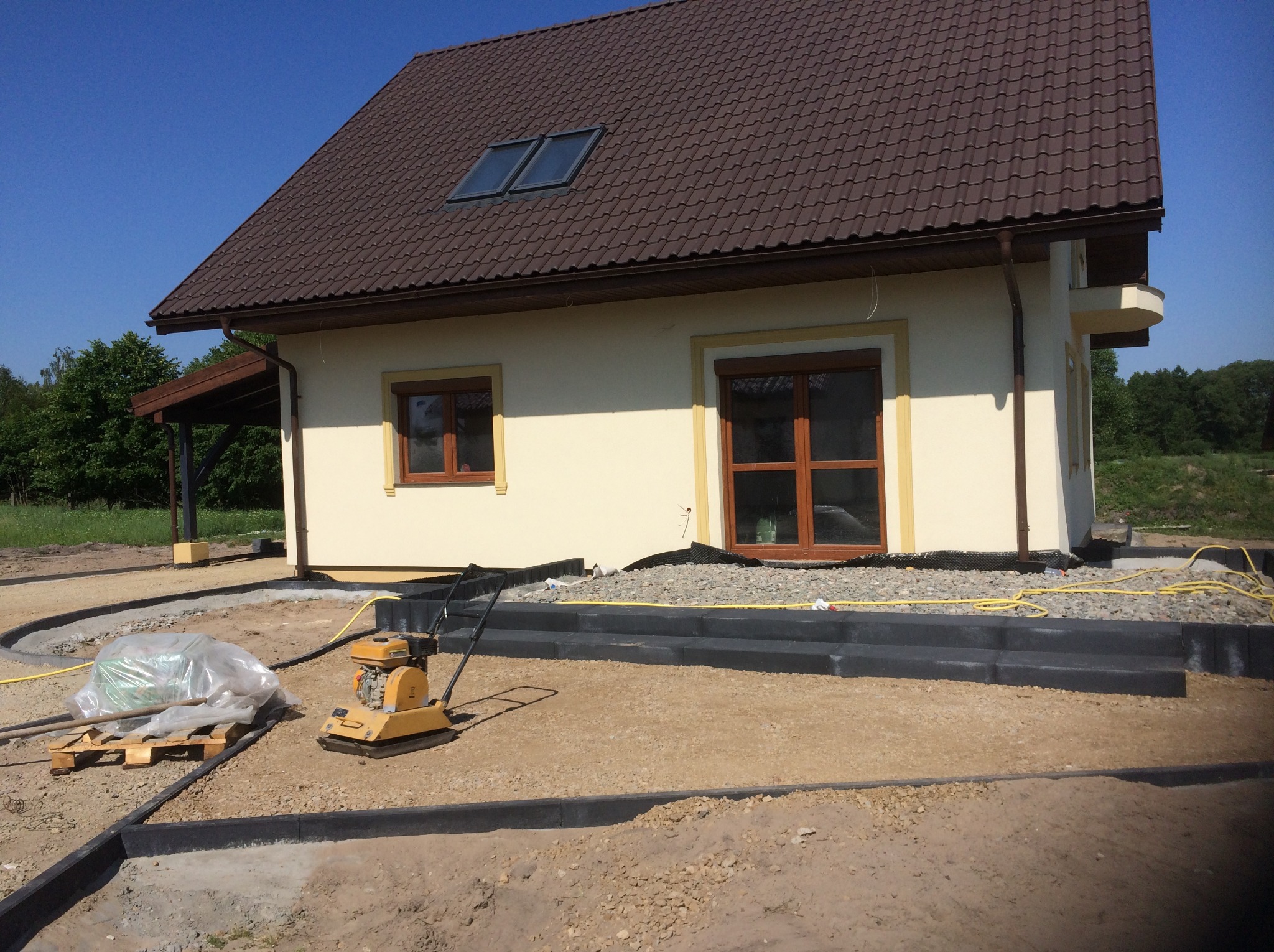 Żółty zagęszczarka gruntu stoi na piasku obok palety z workiem materiału, widoczne obrzeża posesji i fragment ocieplonego budynku z brązową dachówką.