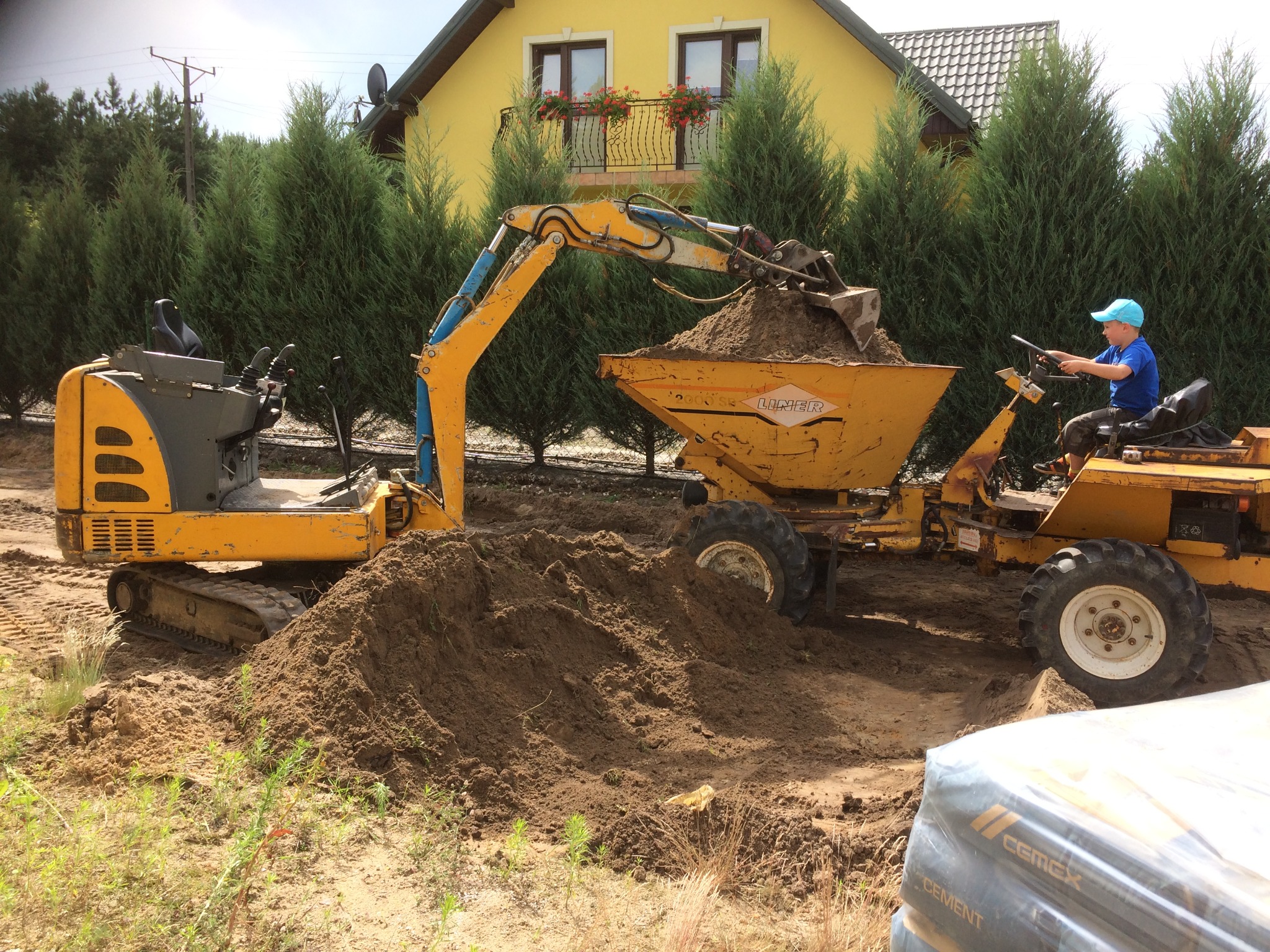 Żółta koparka z łyżką nabierająca ziemię do wywrotki prowadzonej przez chłopca w niebieskiej czapce, na tle domu z balkonem i tujami, widoczny stos worków z cementem.