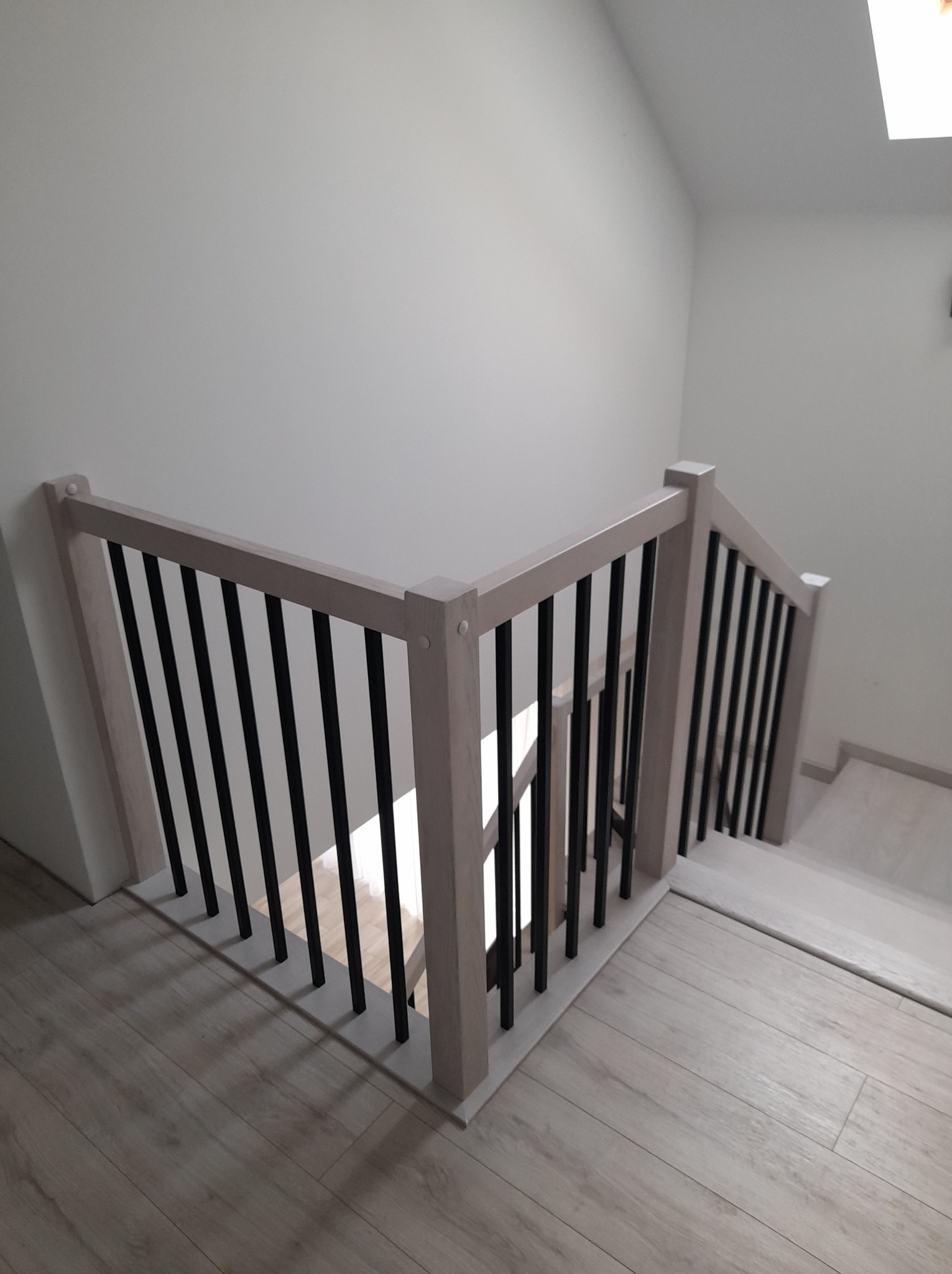 Jasne schody z czarnymi, pionowymi balustradami w nowoczesnym wnętrzu, widok z góry na pierwszy bieg schodów.