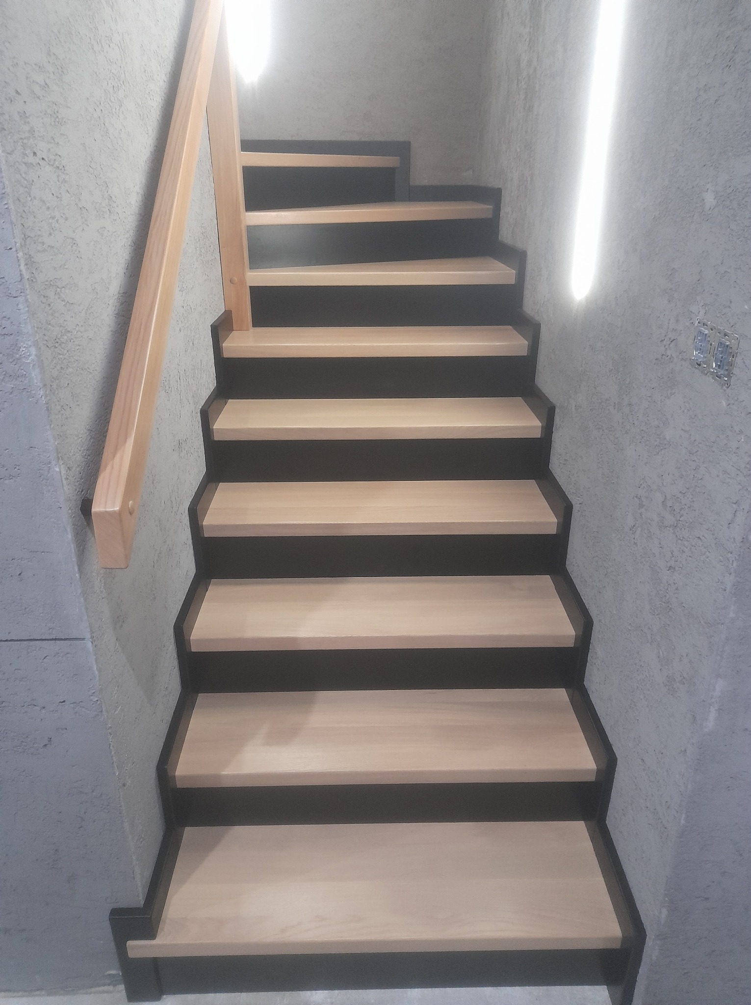Bieg schodów z jasnego drewna z ciemnymi podstopnicami, widoczne z dołu, z drewnianą poręczą po lewej stronie i oświetleniem LED na ścianie po prawej.