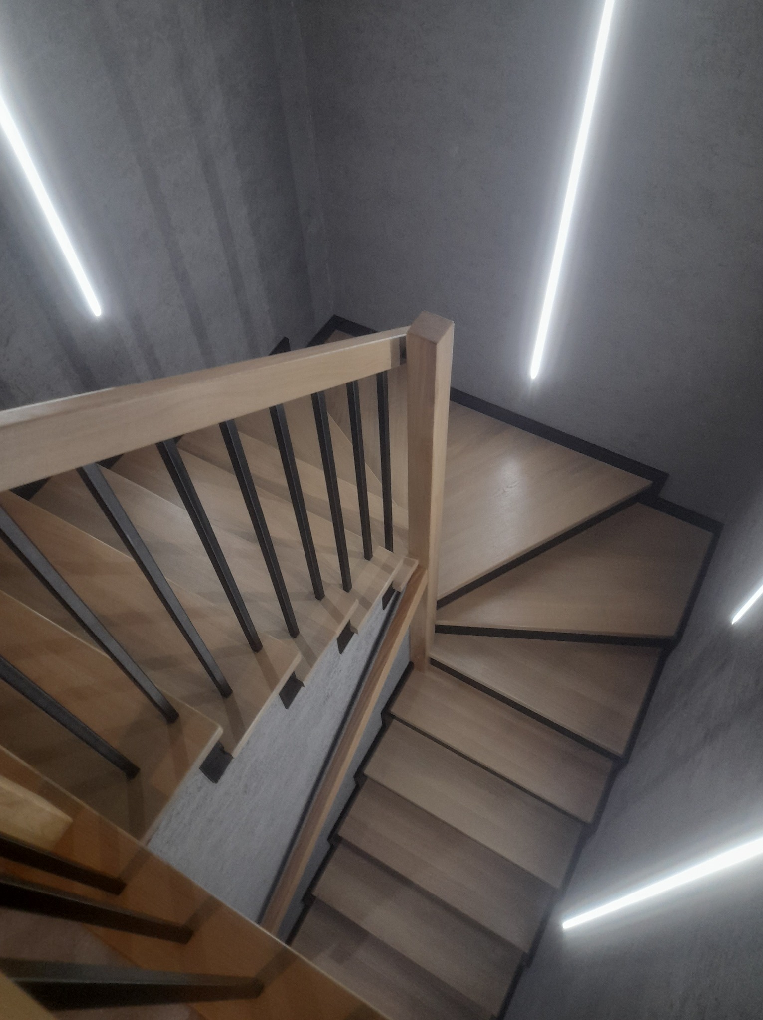 Spiralne schody drewniane z czarnymi balustradami, oświetlone liniowymi lampami LED w minimalistycznym, betonowym wnętrzu, widok z góry.