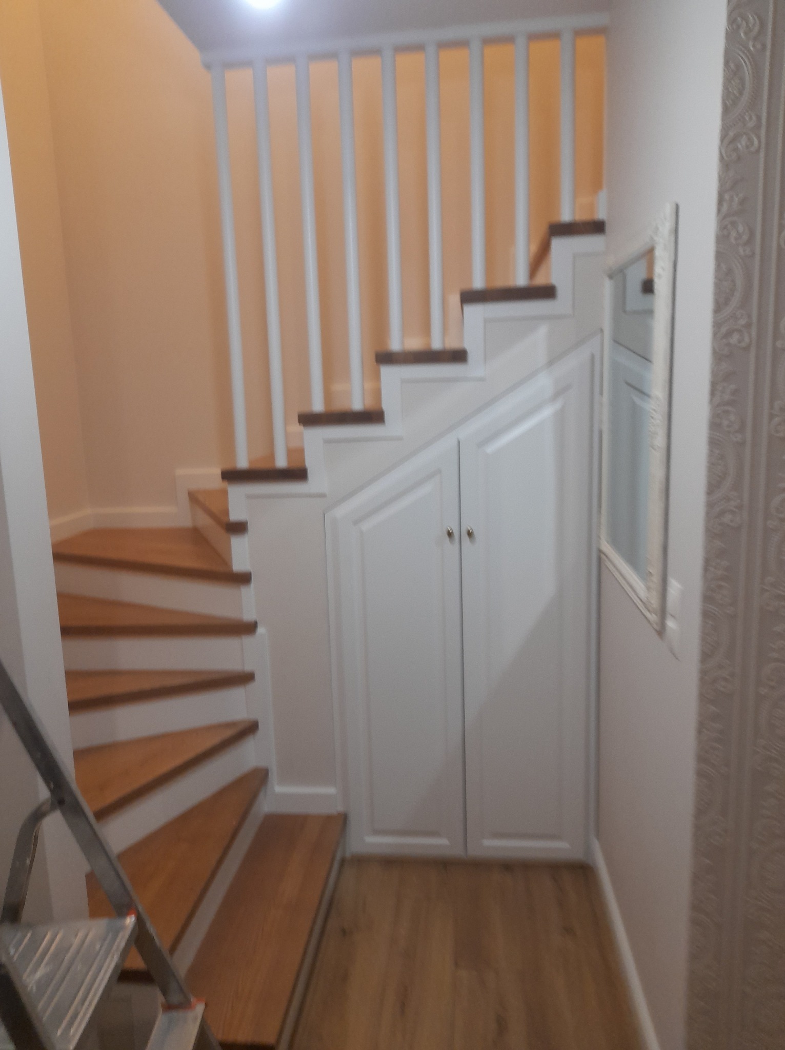 Bieg schodów z drewnianymi stopniami i białą balustradą, zabudowany szafkami pod schodami, widoczne fragment drabiny malarskiej.