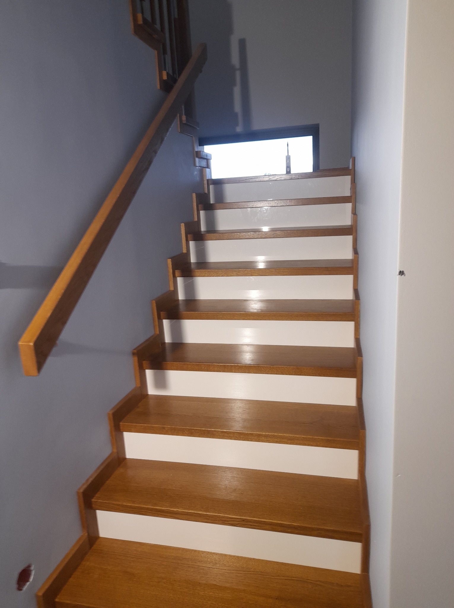 Nowoczesne schody o drewnianych stopniach z białymi podstopnicami i drewnianą poręczą na tle szarej ściany, widoczne okno na podeście.